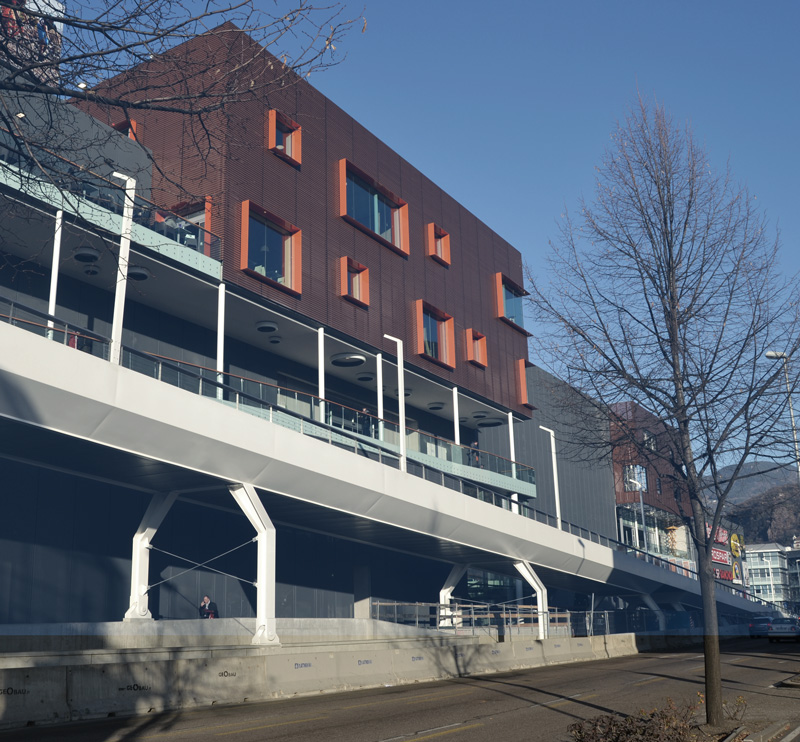 New shopping mall block, exterior view. The ramps take the pedestrian paths directly into the shopping center or to the bridge that crosses the street. The external glazed parapets use the same system as the internal ones with the wooden handrail to give continuity.
